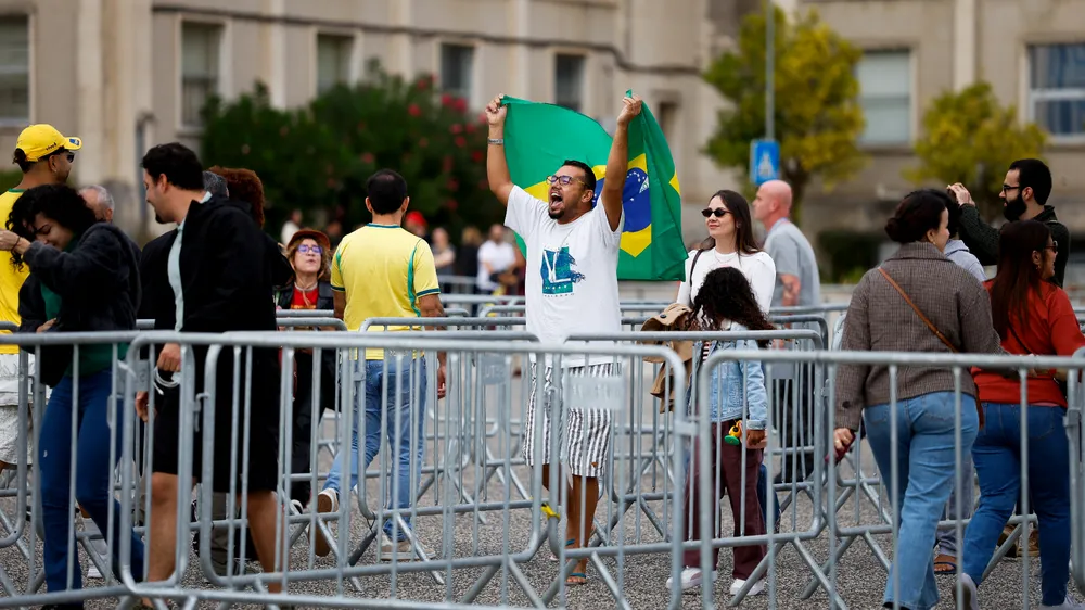 Eleitores aguardam entre grades metálicas, enquanto um homem ergue uma bandeira do Brasil.