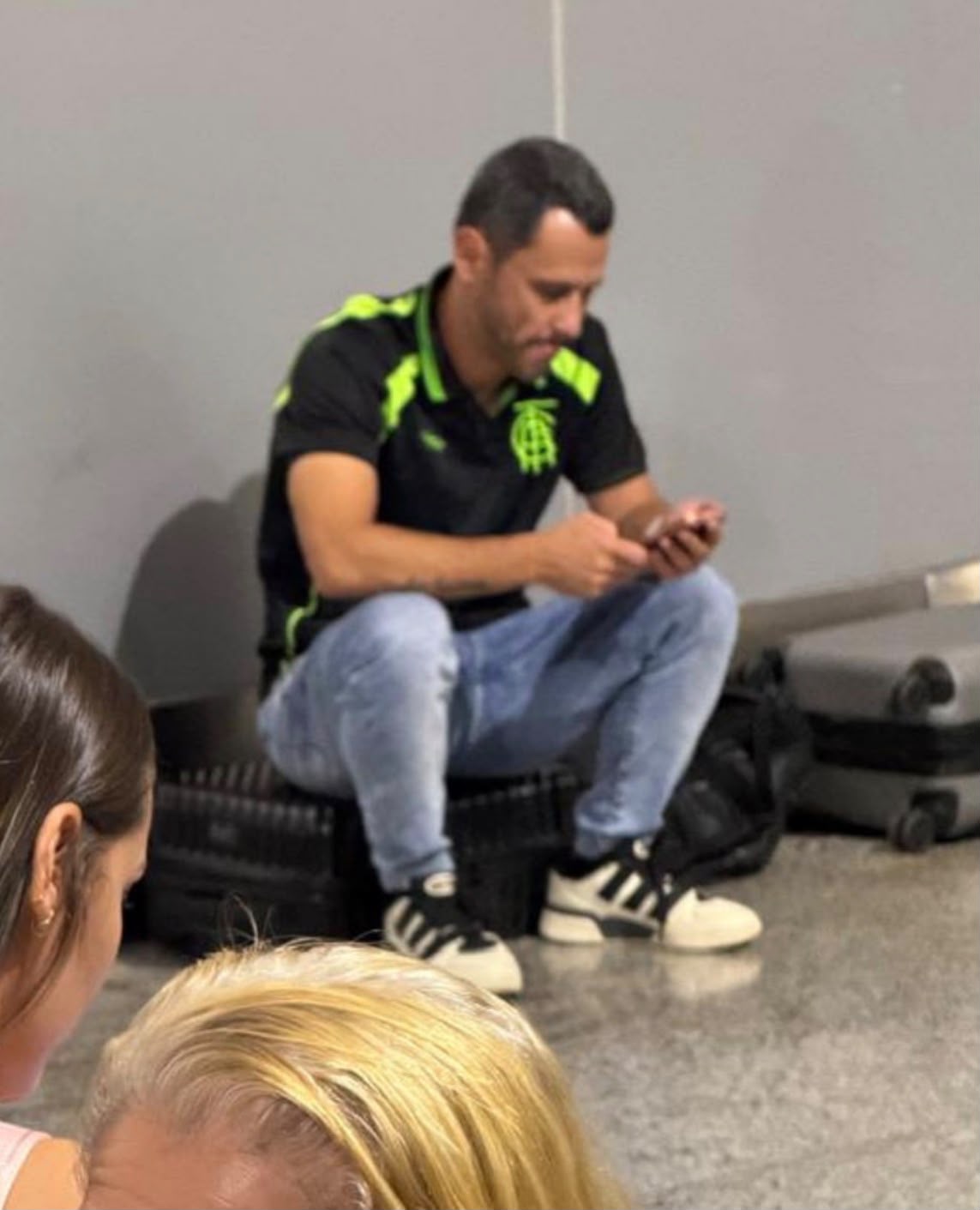 Cleitinho segura um celular, com os joelhos dobrados, junto à parede e às bagagens no aeroporto.