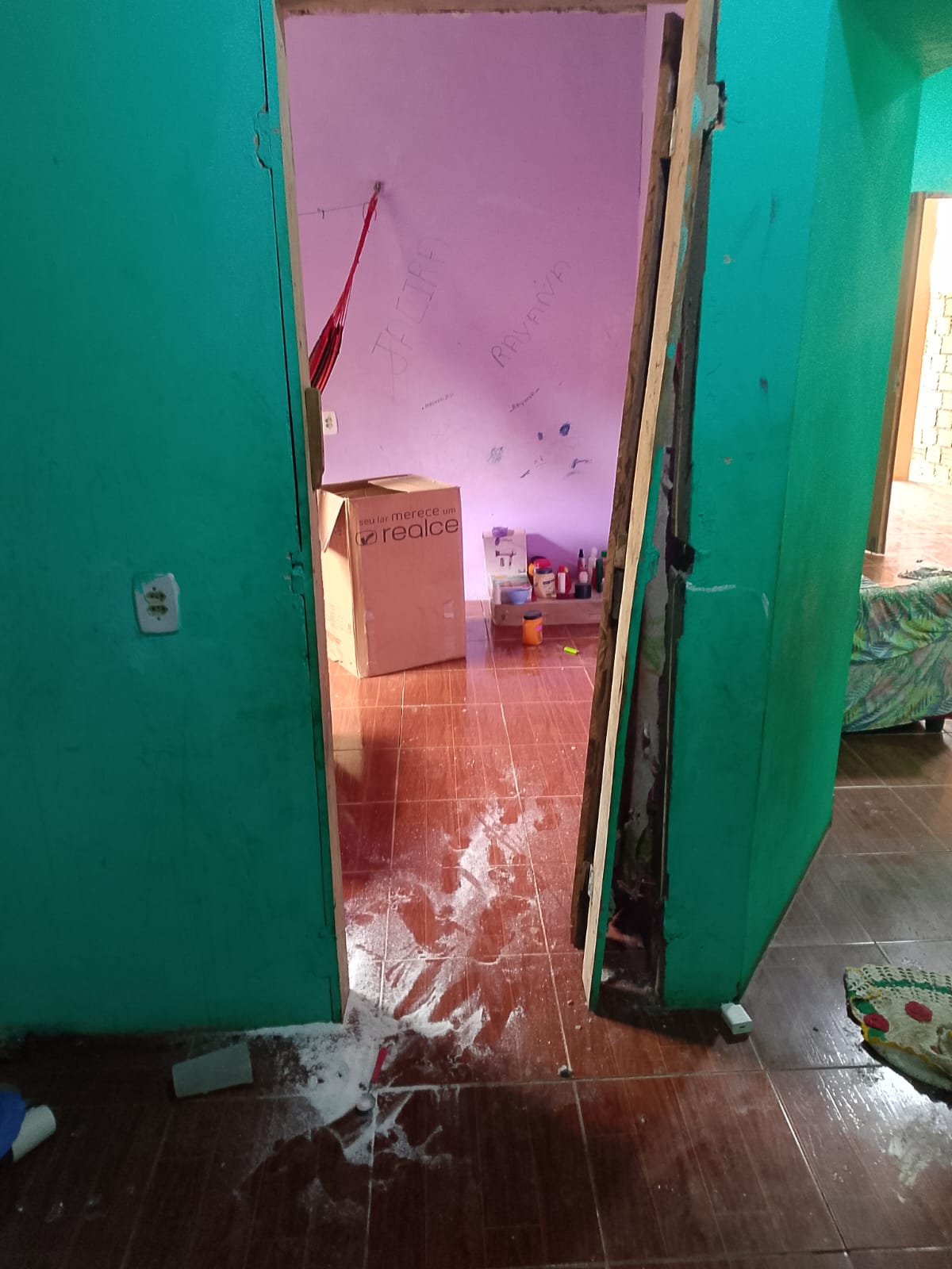 Interior de casa com porta avariada e chão sujo, mostrando danos.